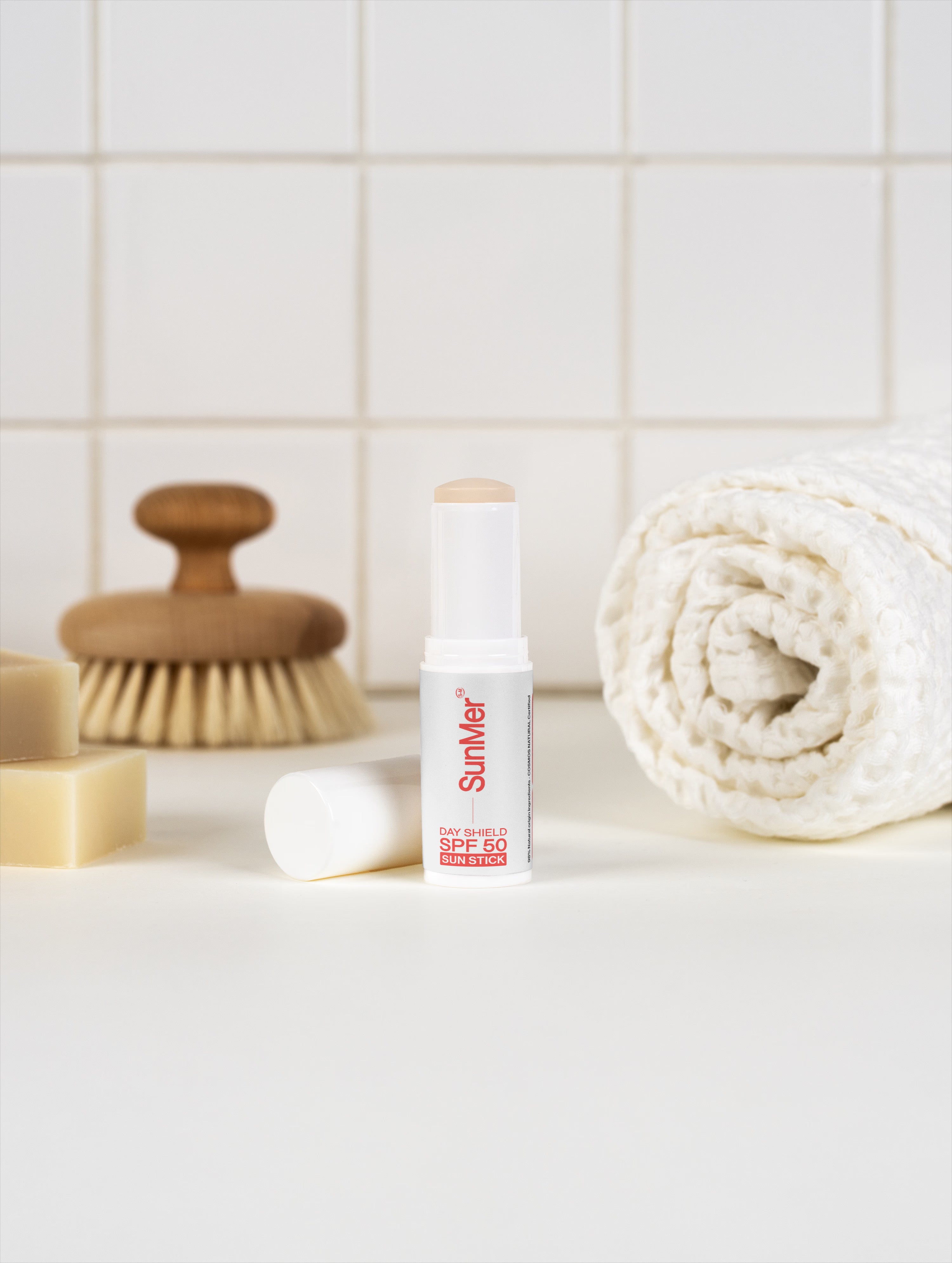 SunMer Day Shield High-Performance SPF50 Sun Stick — COSMOS Natural Certified. SunMer skincare product on a bathroom counter with a towel and brush in the background